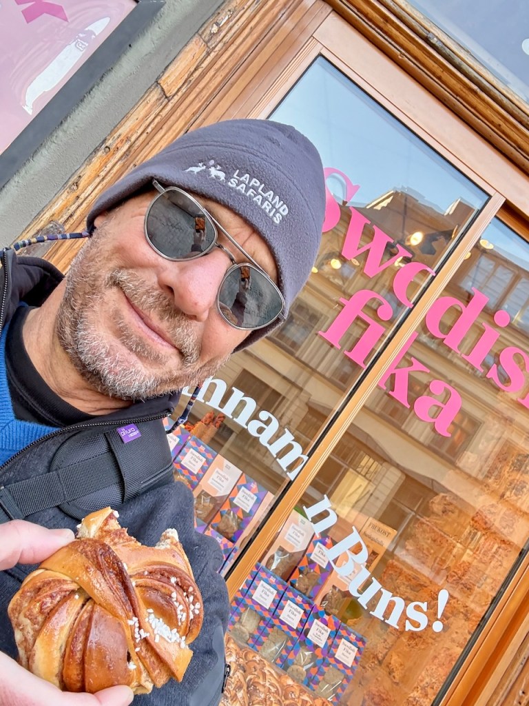 A person wearing sunglasses and a beanie is smiling while holding a cinnamon bun in front of a bakery window with the words 'Swedish Fika' and 'Cinnamon Buns!' displayed.