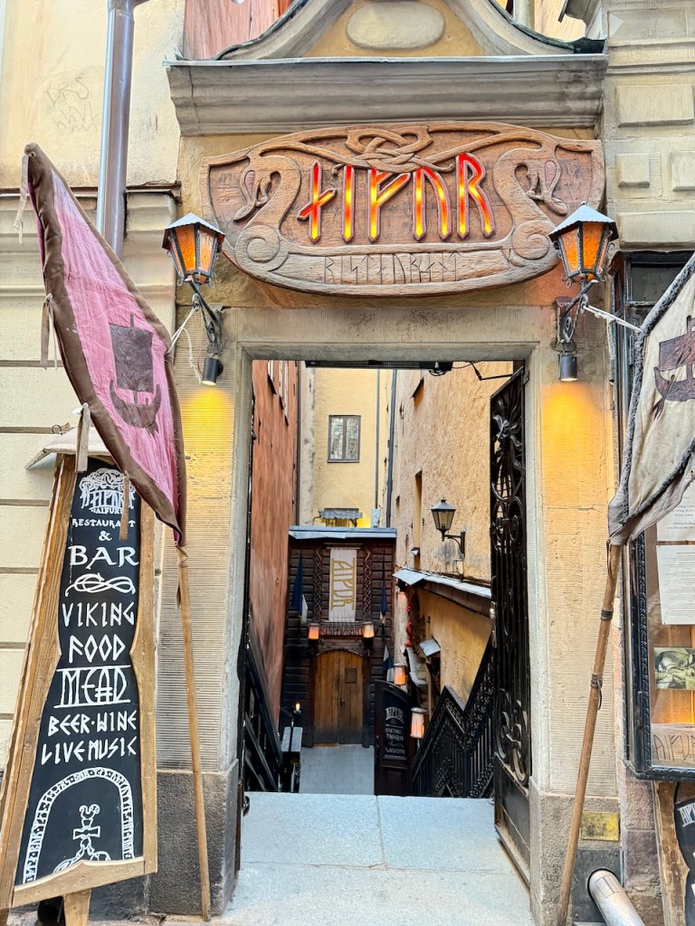 Entrance to a Viking-themed restaurant in Stockholm, featuring a wooden sign with neon letters and a rustic facade. A blackboard menu displays offerings such as Viking food, mead, and live music.