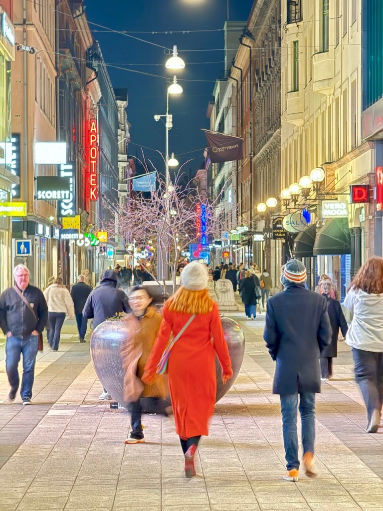 A bustling nighttime street scene in Stockholm, featuring pedestrians walking along a well-lit avenue lined with shops and restaurants. A woman in a bright red coat walks ahead, with various people in winter attire surrounding her.