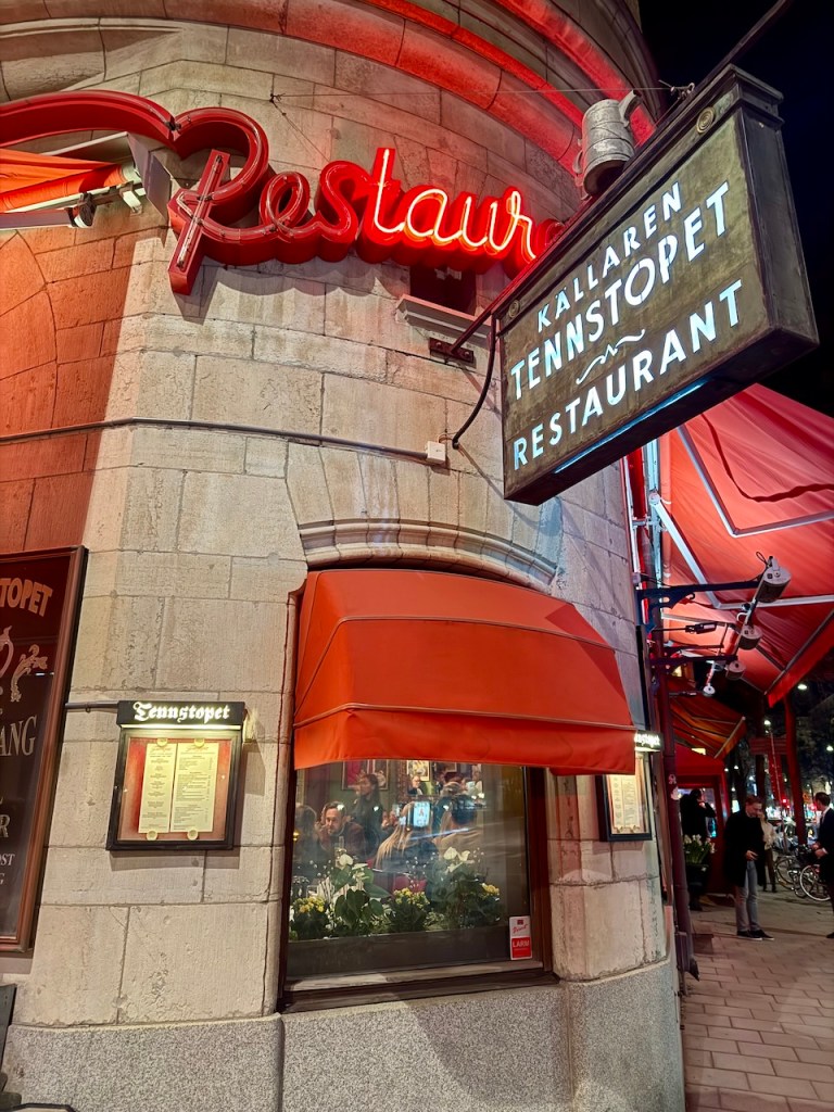 Exterior view of the Tennstopet restaurant in Stockholm at night, featuring a neon red sign and a traditional wooden sign with the restaurant's name.