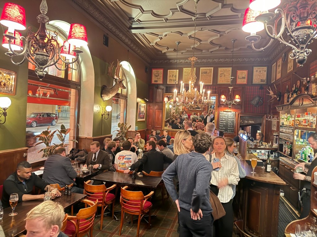 Interior of a vibrant Swedish restaurant and bar with patrons socializing at tables, chandeliers hanging from the ceiling, and decorative elements on the walls.