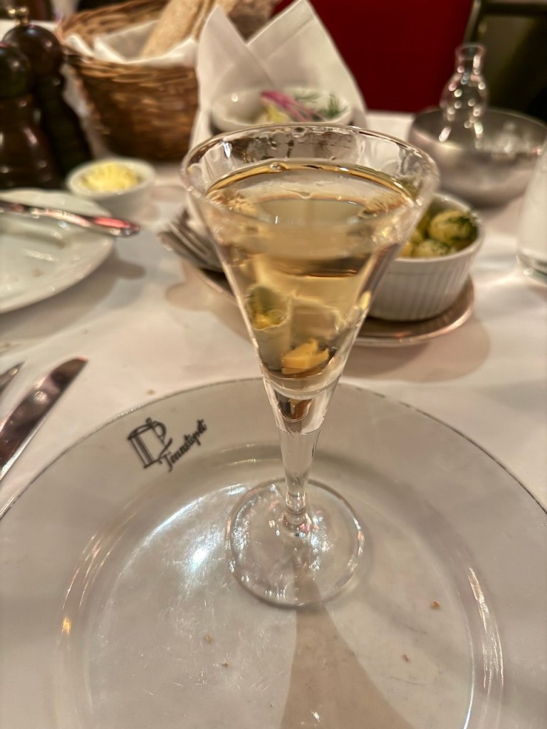 A close-up view of a glass of schnapps served in a tall, slender glass on a plate bearing the logo of the Tennstopet Restaurant, surrounded by various dining items including a basket of bread and small bowls.