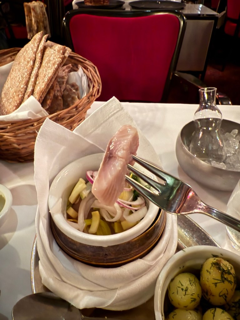 A plate featuring herring served with onions and pickles, alongside a basket of crisp bread and a dish of seasoned olives, in a restaurant setting.