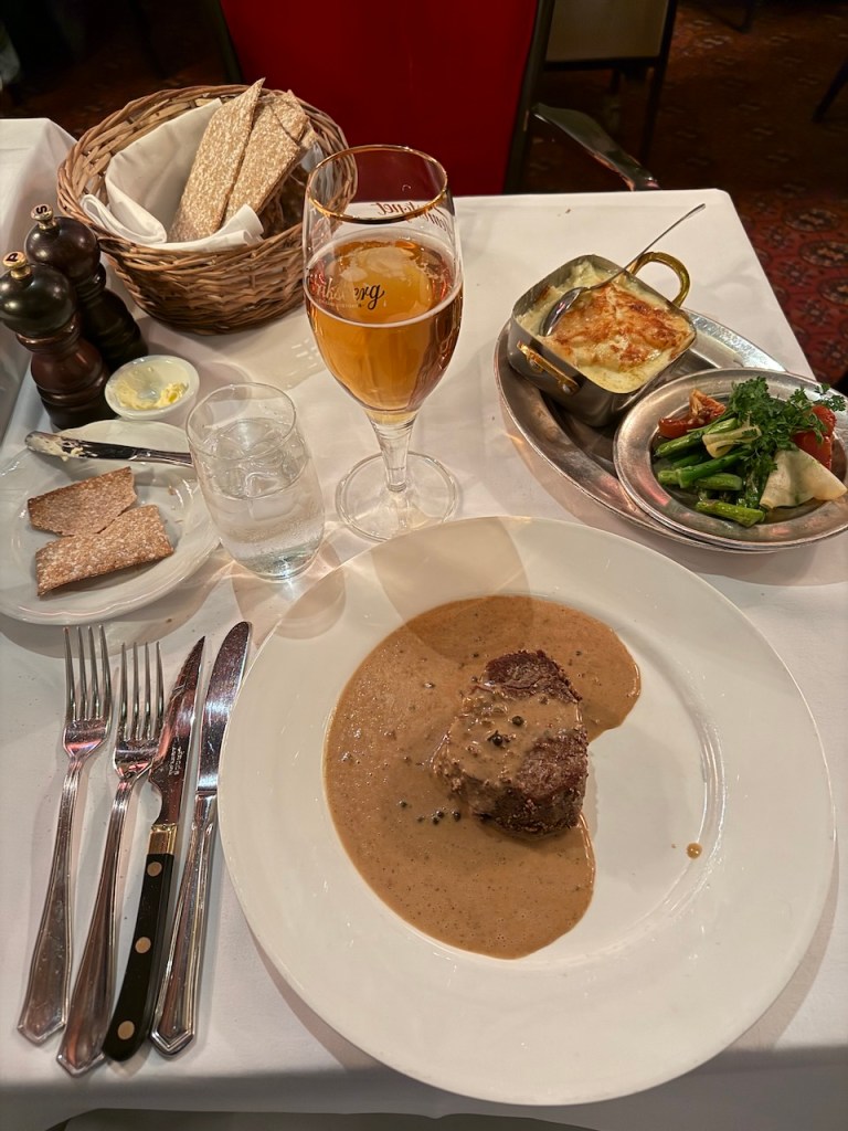 A table setting featuring a plate of traditional Swedish meatballs with gravy, accompanied by a small dish of gratin, mixed vegetables, and a basket of crispbread. A glass of light beer and a glass of water are also present.