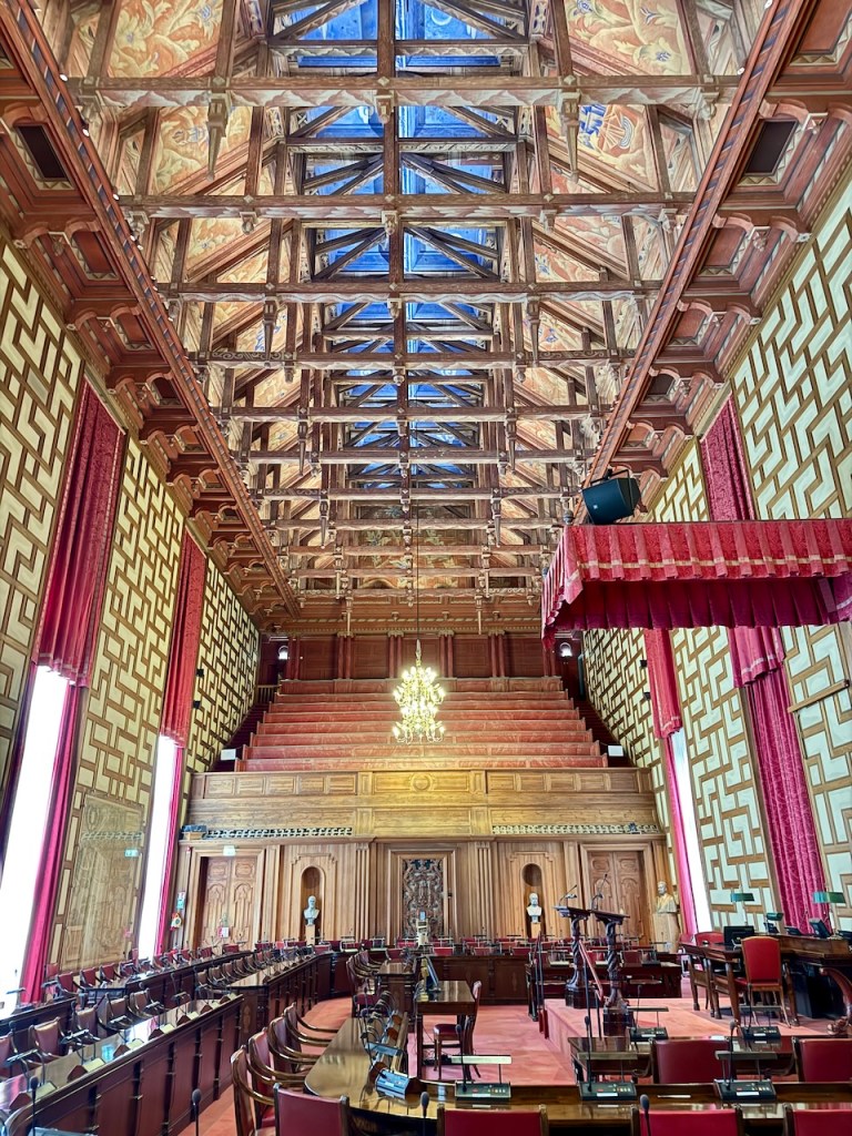 Interior view of a grand legislative chamber featuring ornate wooden beams, red drapes, and a chandelier. The seating arrangement shows rows of desks and chairs, alongside decorative elements on the walls.