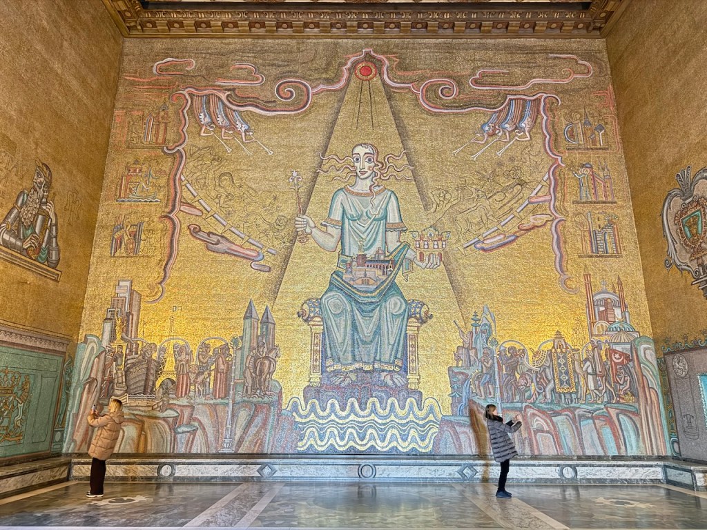 A grand mural depicting a female figure seated, surrounded by various historical symbols and architecture, within the Stockholm City Hall.
