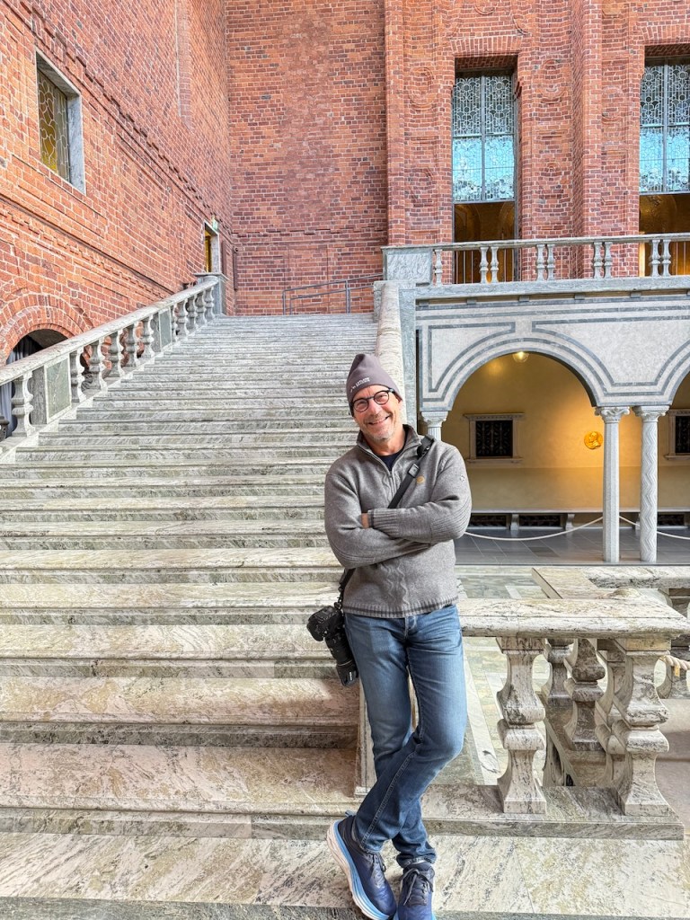 A person smiling while standing on a marble staircase in a brick building, holding a camera.