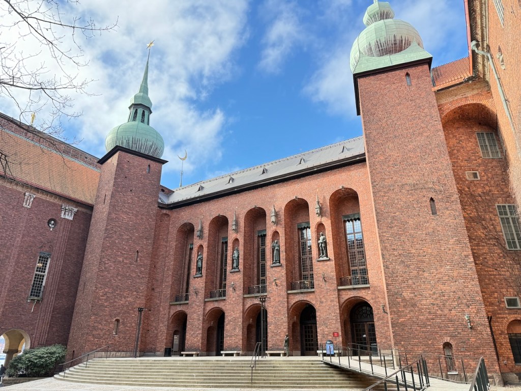 Exterior view of Stockholm City Hall with red brick architecture and green domed towers against a blue sky.