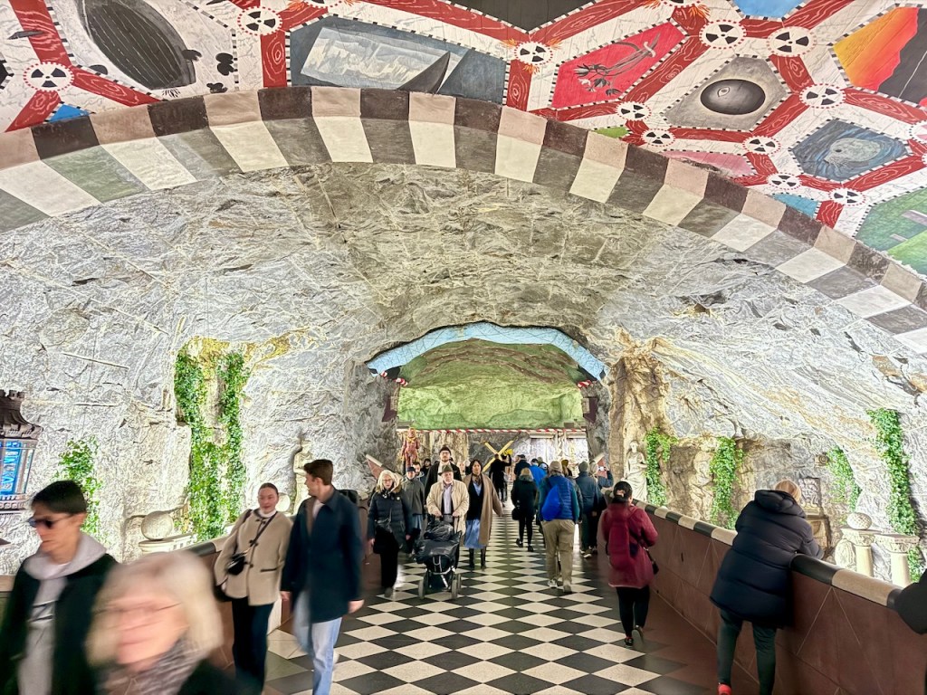 A crowded underground metro station in Stockholm, featuring a stone wall adorned with greenery and colorful murals on the ceiling, with people walking on a black and white checkered floor.