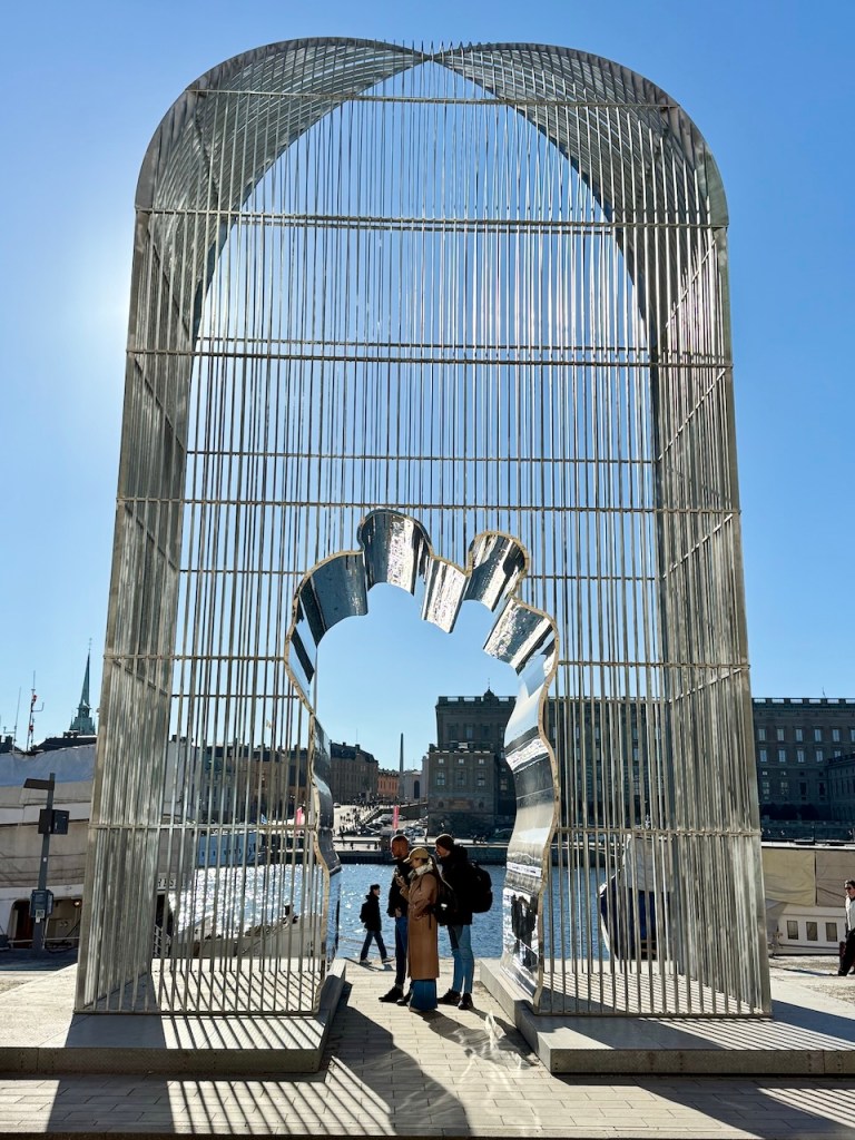 An artistic metallic arch sculpture with a reflective surface, framing a view of a waterfront in Stockholm. Three people stand underneath the arch, enjoying the view, while historical buildings are visible in the background against a clear blue sky.