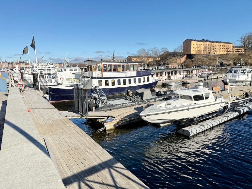 A scenic view of a marina in Stockholm, featuring various boats docked along a wooden pier under a clear blue sky.