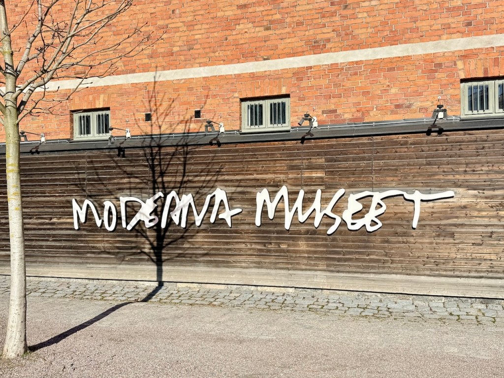 Signage for the Moderna Museet, a modern art museum, on a wooden wall with a brick background.