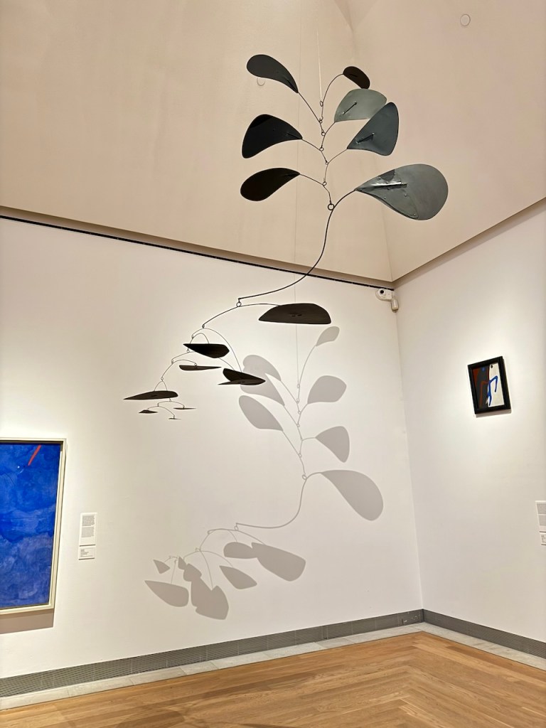 A mobile sculpture with dark, leaf-shaped components hangs from the ceiling in an art gallery, casting intricate shadows on the wall. The scene includes a vibrant blue painting on the left and a small framed artwork on the right, with wooden flooring beneath.