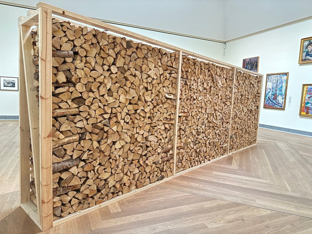 A wooden sculpture made of neatly stacked firewood, displayed in a contemporary art gallery with other artworks visible in the background.