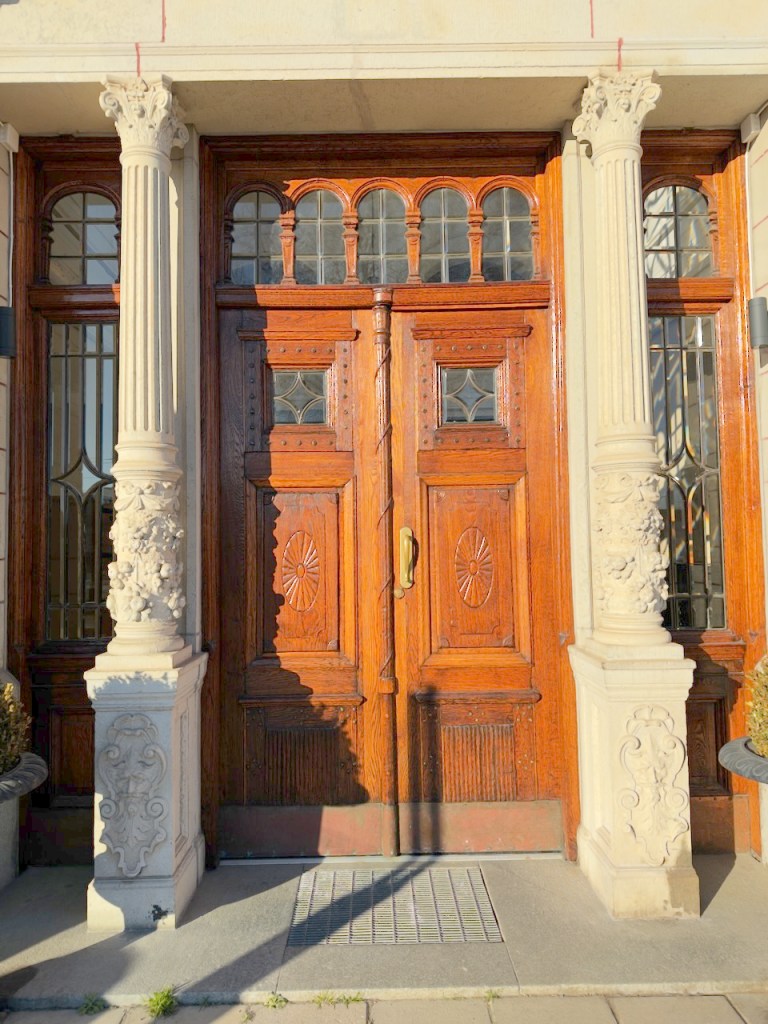 Close-up of an ornate wooden double door with intricate carvings and glass panels, flanked by decorative stone columns.