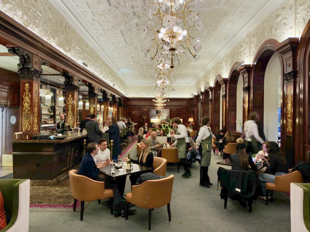 Interior view of a stylish restaurant with elegant decor, featuring customers seated at tables, a bar area with staff, and ornate chandeliers illuminating the space.