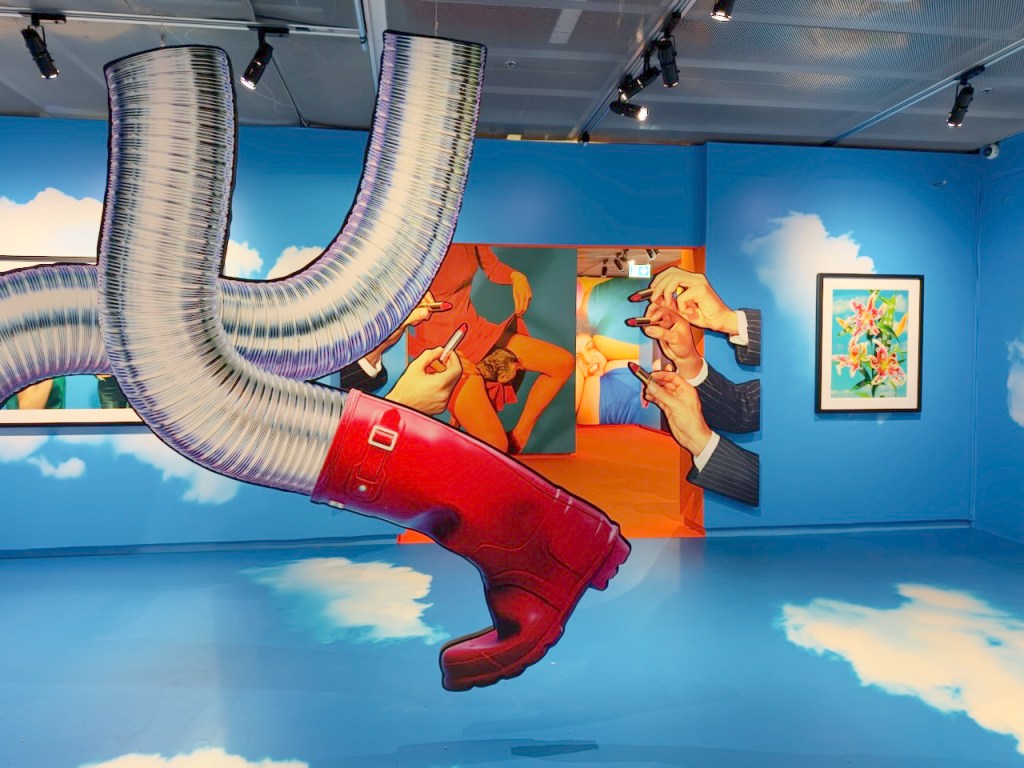 An art installation in a gallery featuring a large red rubber boot and silver flexible ducts against a blue sky backdrop, with colorful art pieces, including hands interacting with a figure, visible on the walls.
