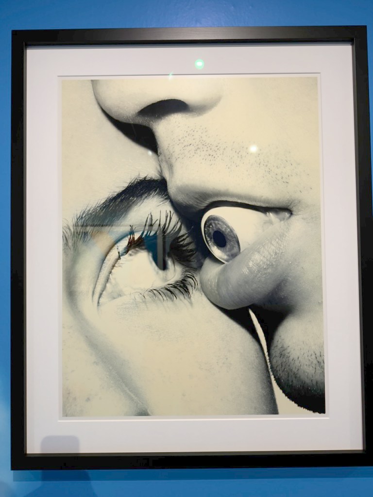 Close-up artwork featuring two human facial features, including lips and an eye, with a finger positioned near the eye. The image is framed against a blue background.
