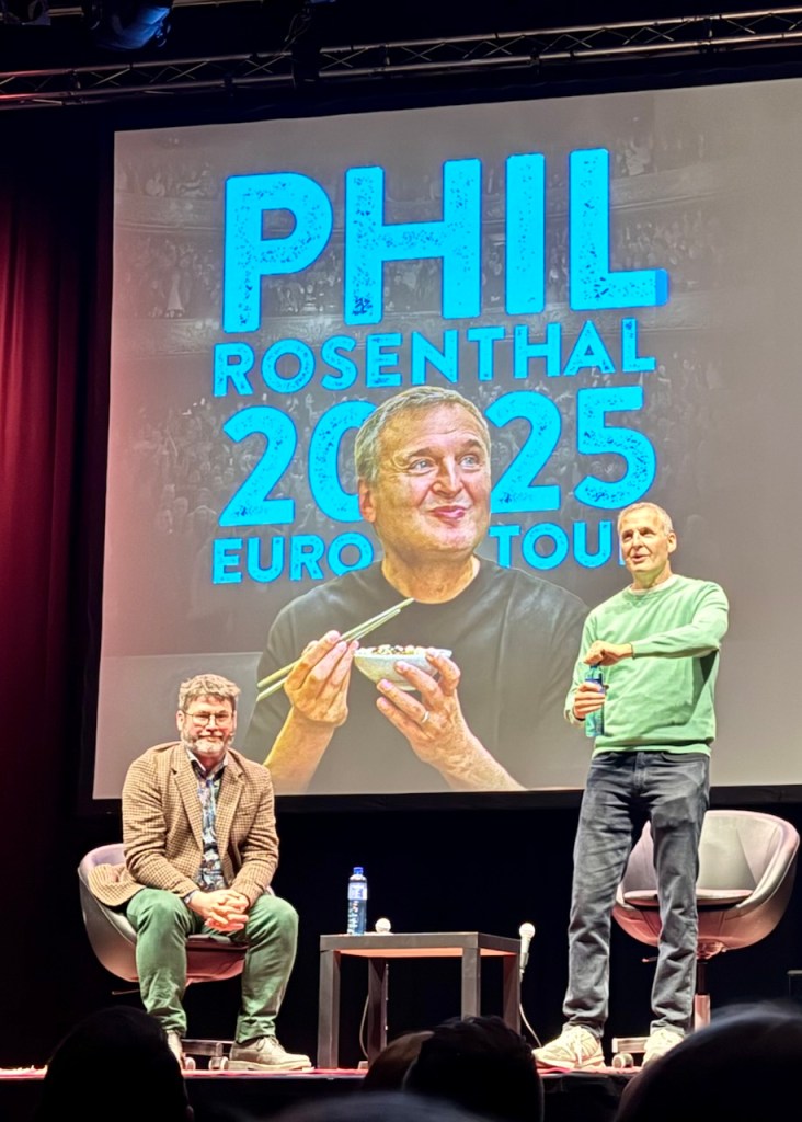 Phil Rosenthal speaking on stage during his 2025 European tour presentation, with a backdrop featuring his name and an image of him enjoying food.