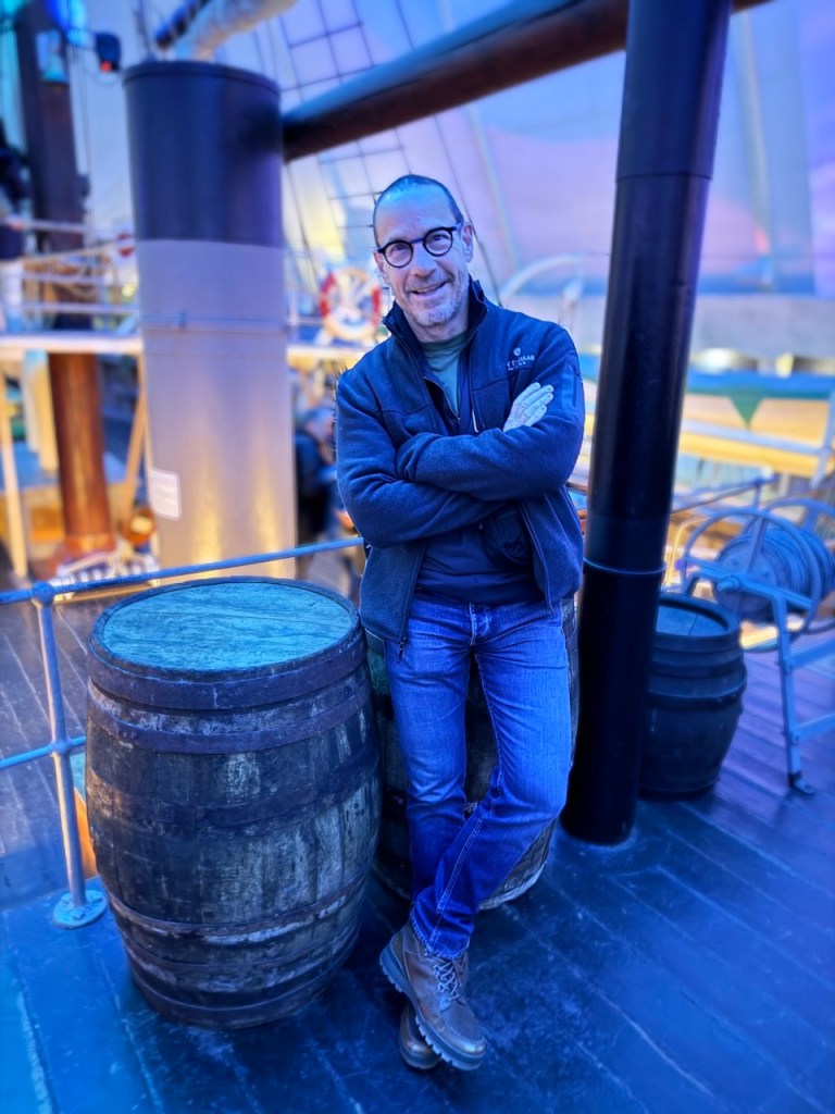 A man standing casually with his arms crossed, next to wooden barrels on the deck of a ship, set in an indoor area with a warm ambiance.