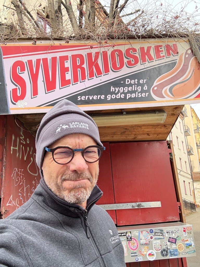 A man standing in front of the closed Syverkiosken hot dog stand in Oslo, with a sign featuring the name and description in Norwegian.