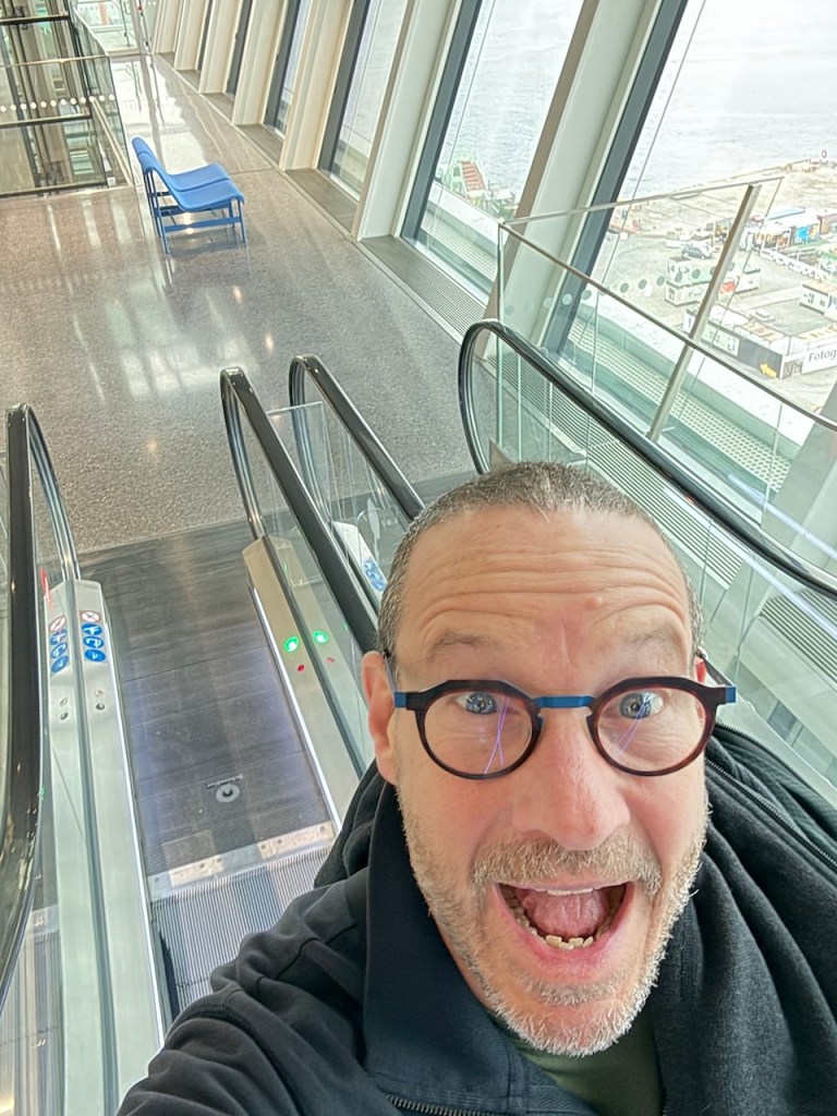 A person making a joyful expression while taking a selfie on an escalator in a modern building, with windows showcasing a view of the harbor.