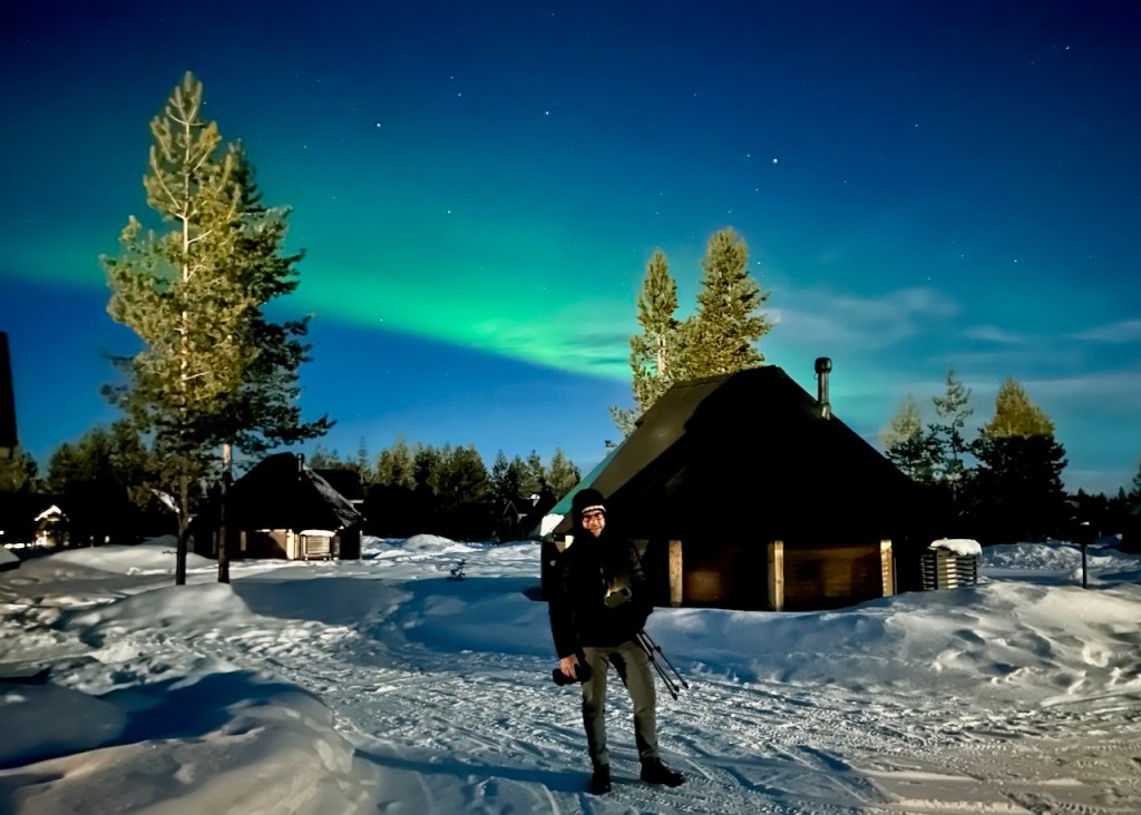 A person standing in a snowy landscape with green auroras lighting up the night sky above, and traditional cabins in the background.