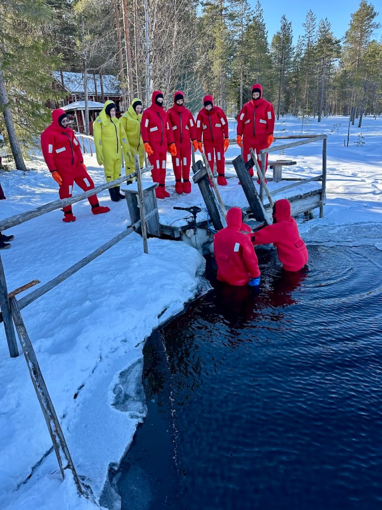 Group of individuals wearing red and yellow insulated wet suits preparing to float in a hole cut into a frozen lake, surrounded by a snowy landscape and pine trees.