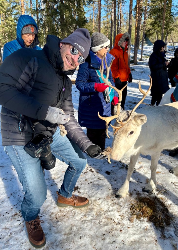 A person smiling and feeding a reindeer in a snowy forest, surrounded by others in winter clothing.