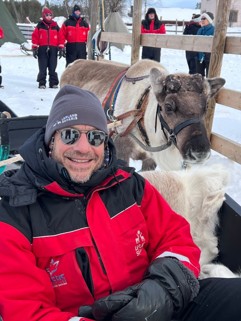 A person wearing sunglasses and a winter jacket smiles while seated next to a reindeer in a snowy environment, with several people in red jackets in the background. The scene depicts a reindeer ride experience in Finnish Lapland.