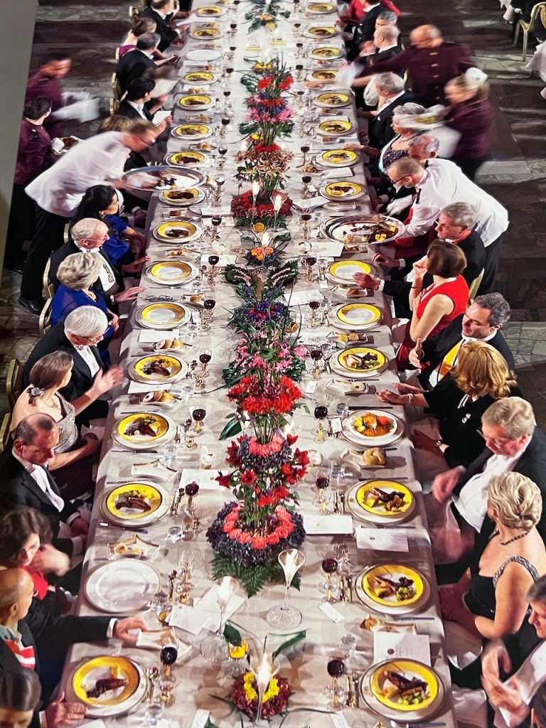 A long banquet table elegantly set for a formal dinner, adorned with floral centerpieces and plates of food. Guests dressed in formal attire are seated on both sides, with servers attending to the meal.