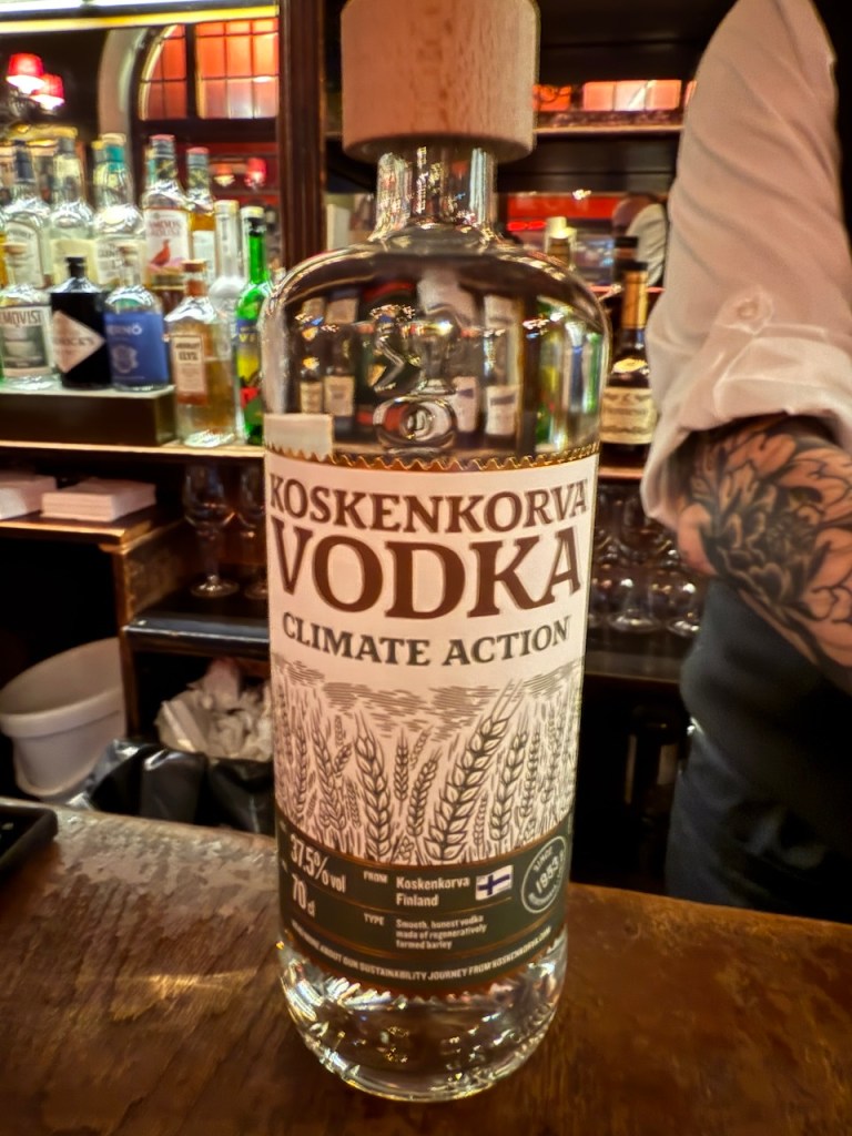 A close-up of a bottle of Koskenkorva Vodka labeled 'Climate Action', sitting on a bar counter with various bottles in the background.