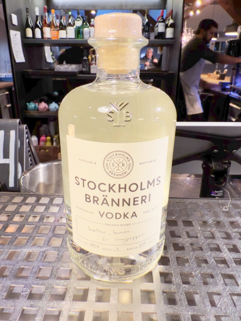 A bottle of Stockholm Bränneri vodka displayed on a metal surface, with the backdrop showing various bottles and a bartender in action.