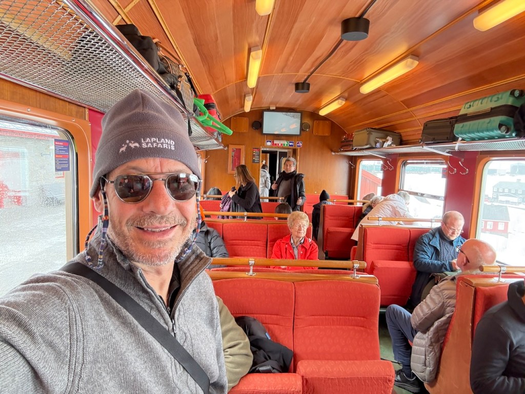 Interior of a train carriage featuring red seats and passengers, with one man in the foreground wearing sunglasses and a beanie.