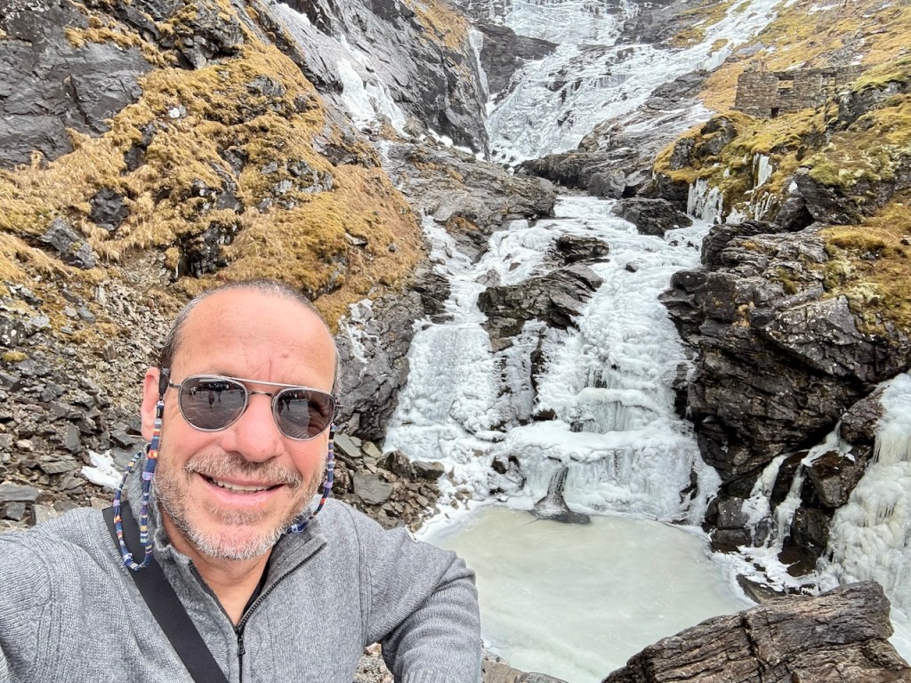 A person taking a selfie in front of a frozen waterfall, surrounded by rocky terrain and patches of grass.