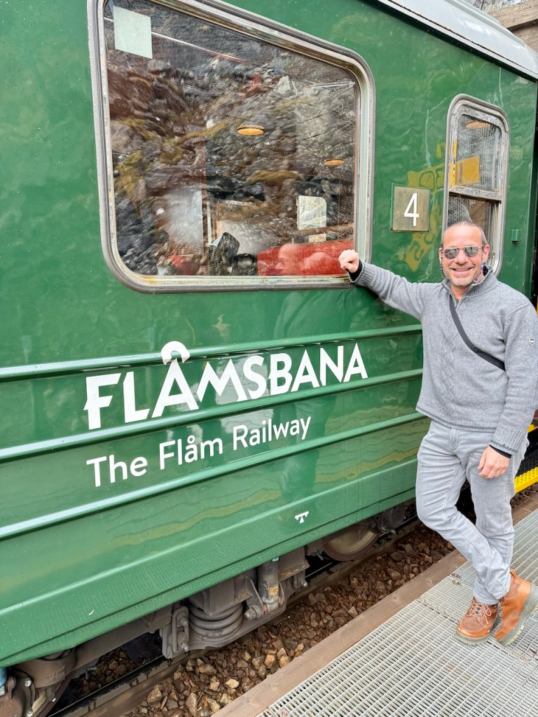 A person standing beside a green train of the Flåm Railway, featuring its name prominently displayed on the side.