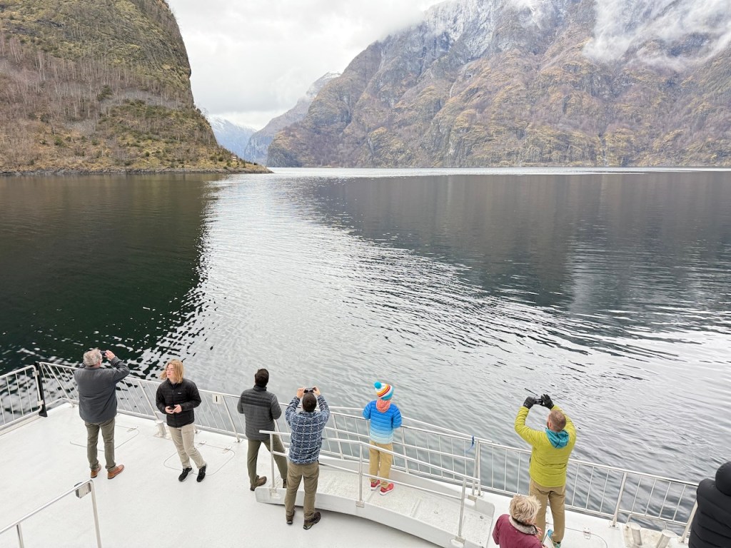Tourists taking photos on a boat cruise in a fjord, surrounded by steep mountains and calm waters.