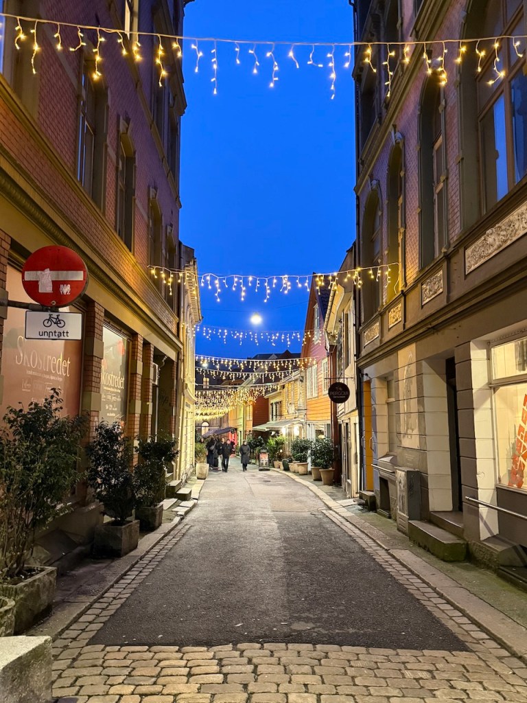 A charming street in Bergen, Norway, adorned with string lights, showcasing colorful buildings and bustling activity under an evening sky.