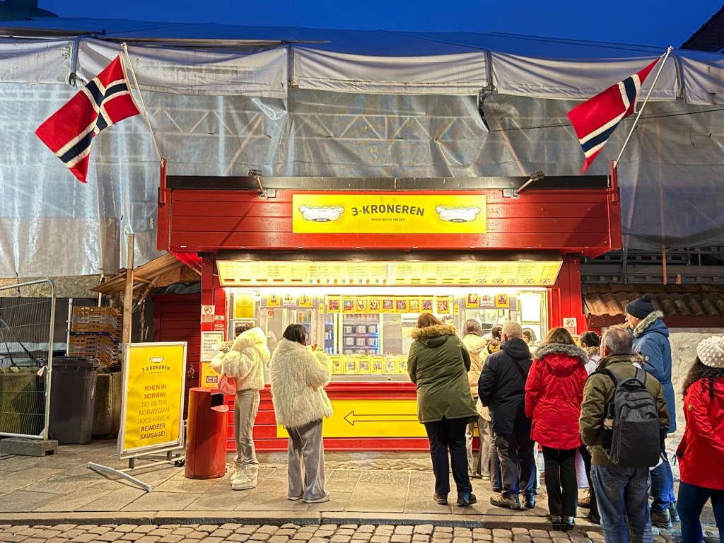 A food stall named '3-Kroneren' in Norway, featuring a bright red exterior with a menu displayed. Patrons wearing winter clothing are lining up to order, while two Norwegian flags are visible above the stall. The scene is set at night, illuminated by warm lights.