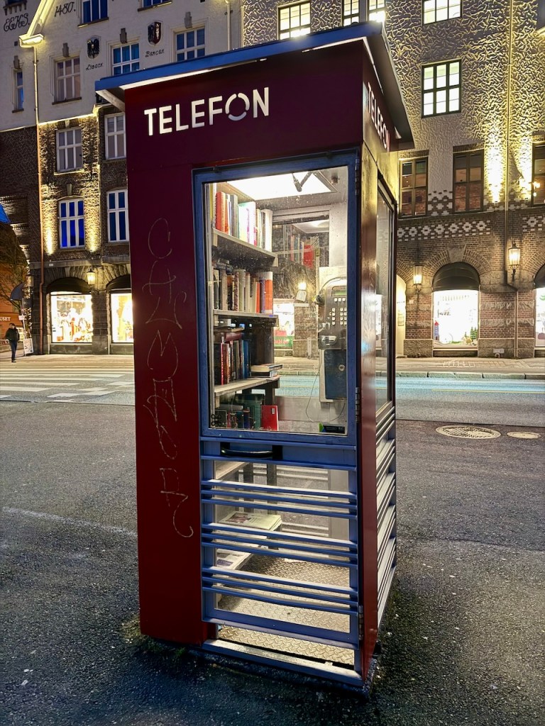 A red telephone booth transformed into a small public library, filled with books, illuminated at night.