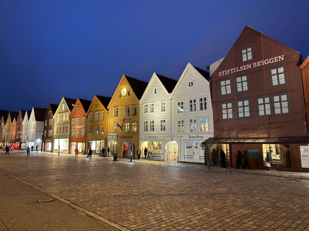 Colorful wooden buildings line a cobblestone street in Bergen, Norway, illuminated by evening lights.