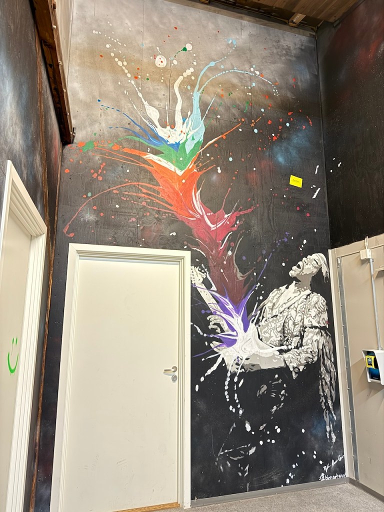 A colorful mural depicting a dynamic figure releasing vibrant splashes of paint, set against a dark background with a door visible on the left.