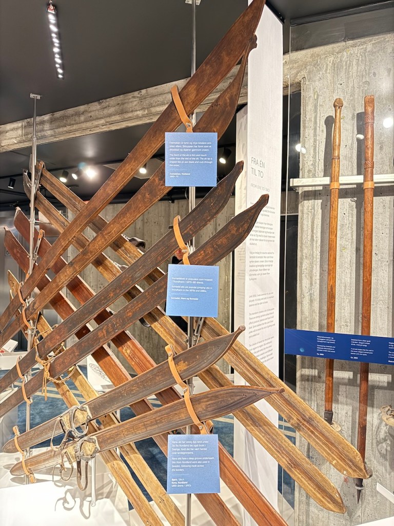A display of traditional wooden skis and ski poles in a museum, featuring various designs and a historical context, with informative plaques in Norwegian.