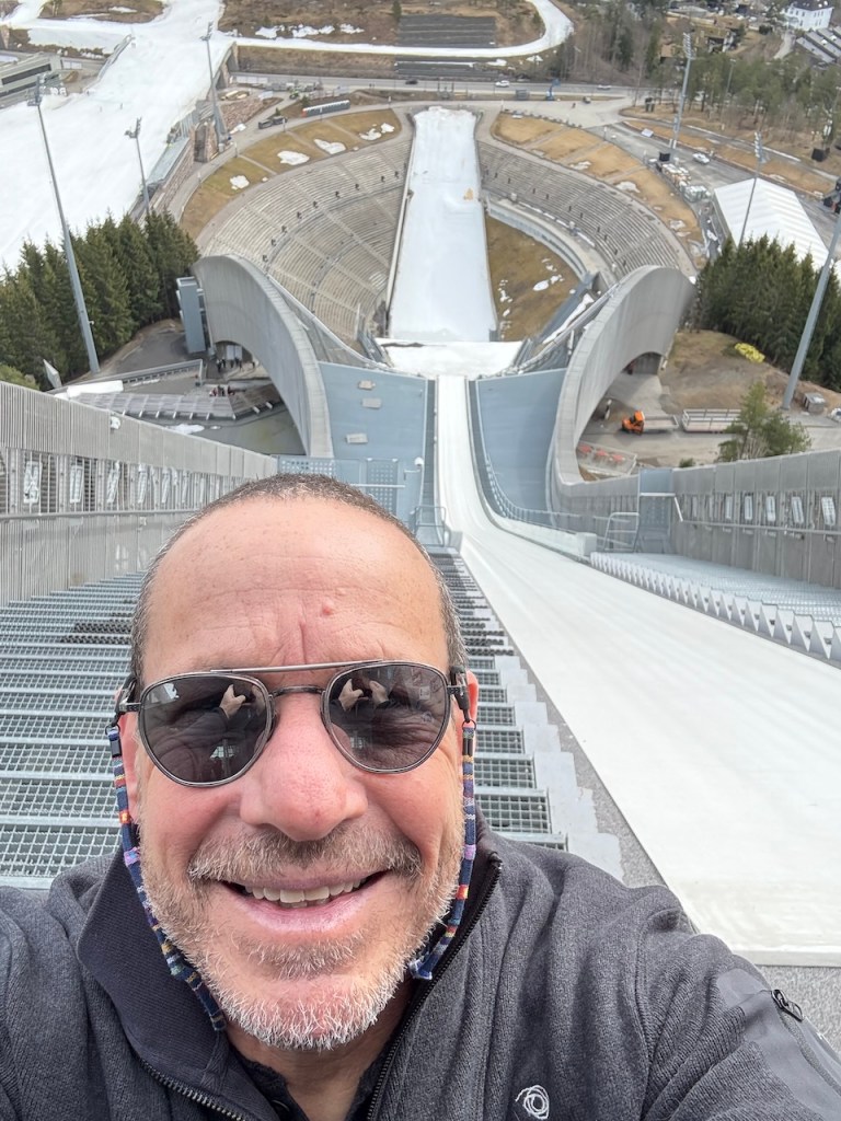 A person taking a selfie from the top of a ski jump with an expansive view of the jump's landing area and surrounding landscape in the background.