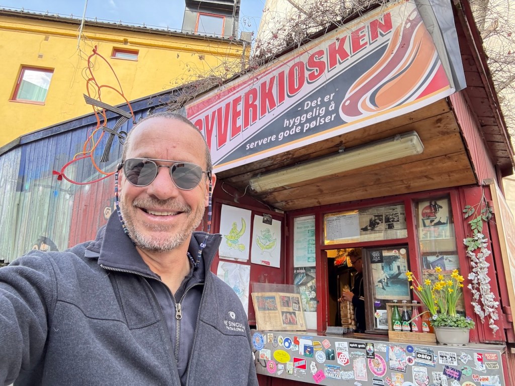 A man stands in front of the Syverkiosken hot dog kiosk, smiling and wearing sunglasses. The kiosk features a colorful sign and is decorated with various stickers, while spring flowers are placed on the counter.