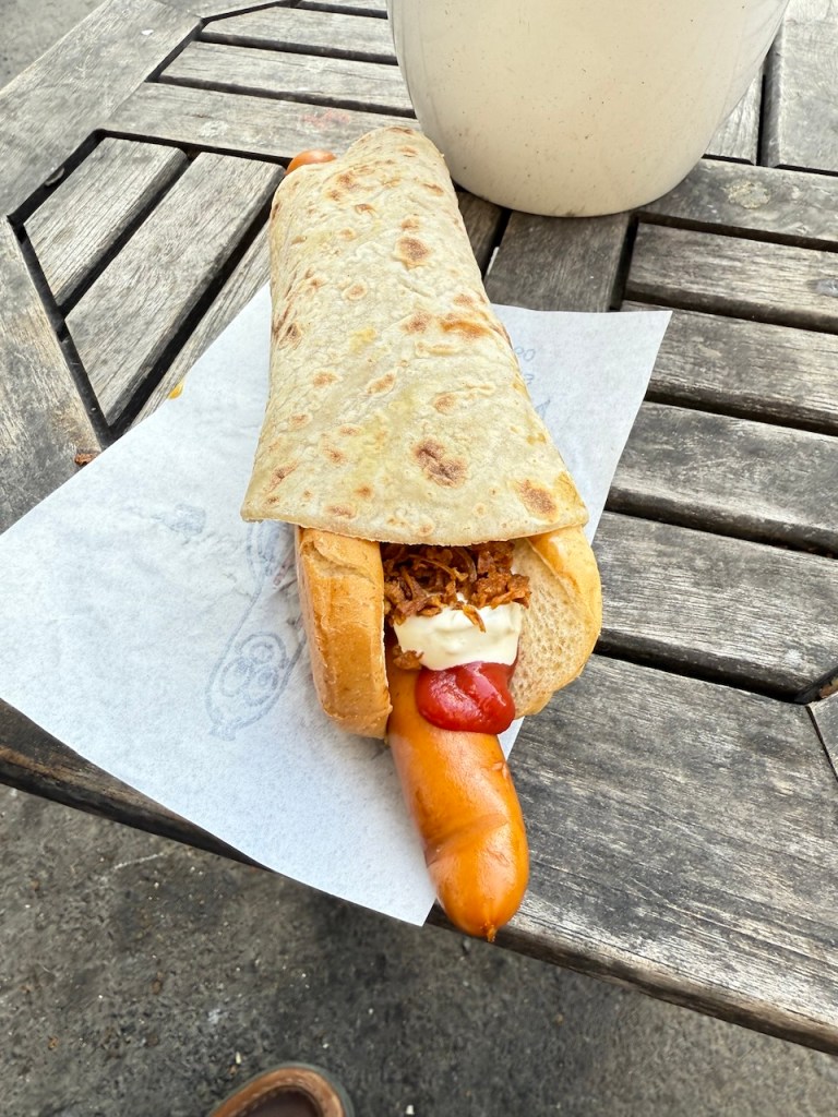 A spicy hot dog wrapped in sausage bread and traditional potato flatbread, topped with homemade ketchup, mustard, jalapeño salad, and crispy fried onions, resting on a wooden table.