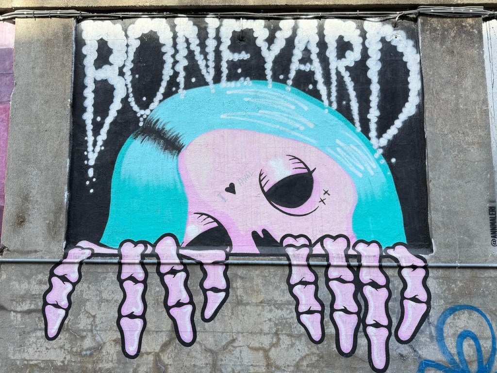 Colorful street art featuring a skull with blue hair and skeletal hands, with the word 'BONEYARD' captioned above in a playful font.