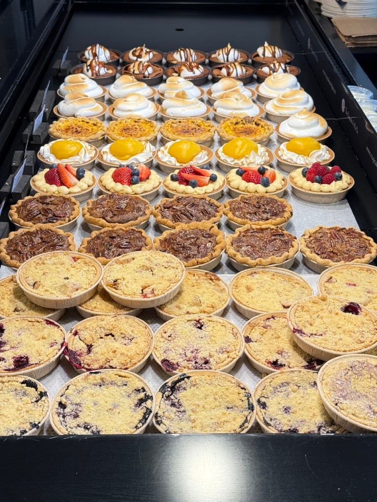 A variety of baked desserts arranged neatly, including fruit tarts topped with strawberries, raspberries, and mango, as well as mini pies with various fillings like pecan and fruit crumble.