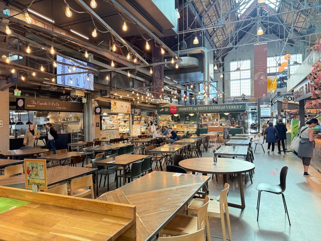 Interior view of Mathallen Oslo, an upscale food court with a modern industrial design featuring wooden tables, string lights, and various food stalls.