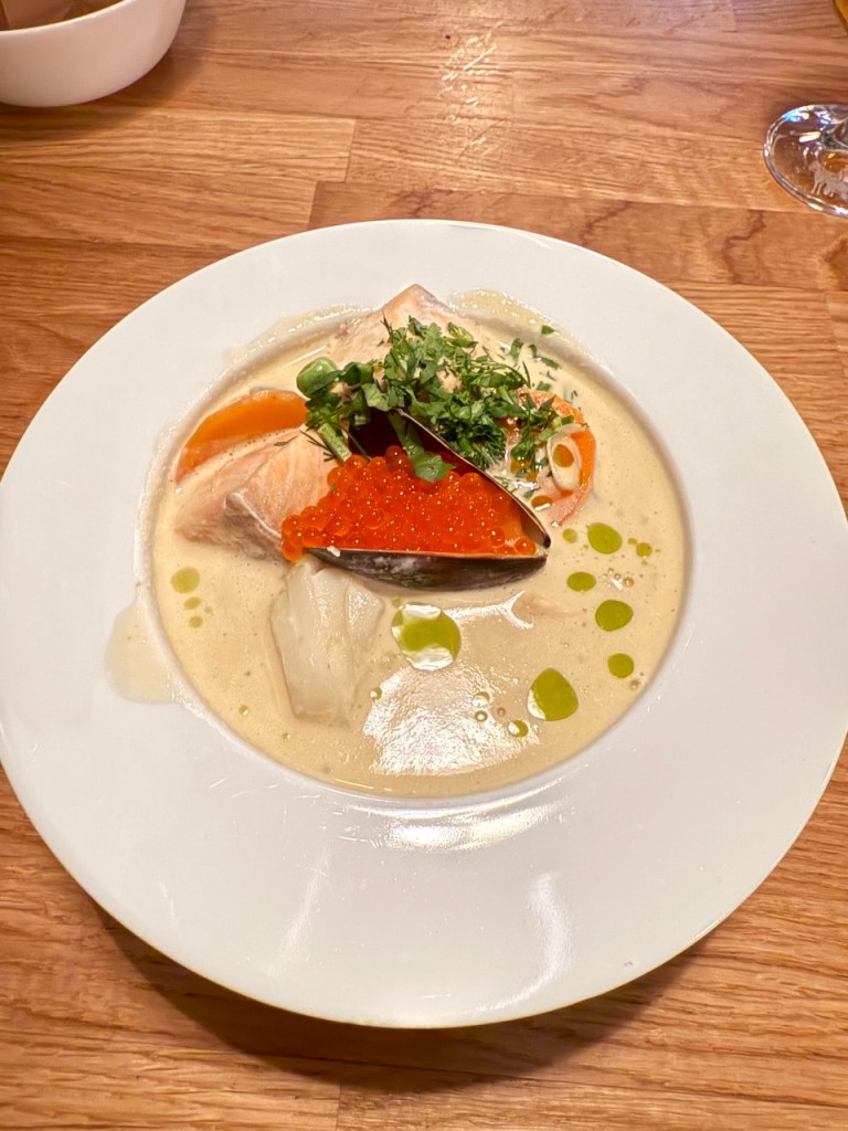 A beautifully presented dish featuring salmon, orange garnishes, and salmon roe on a creamy broth, served on a white plate against a wooden table.